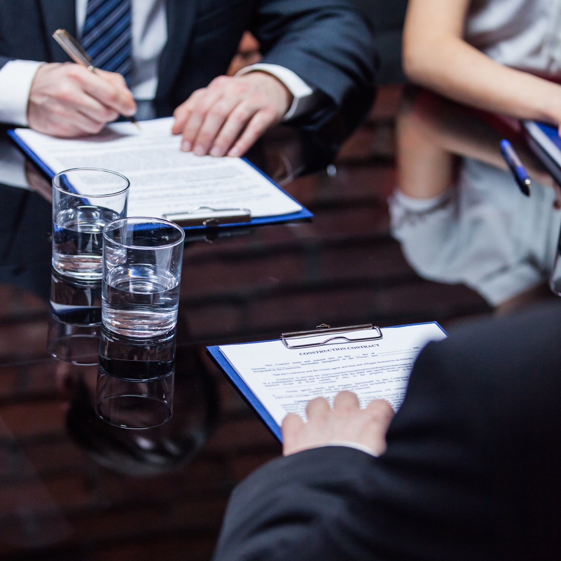Three people discuss the terms of an agreement. They are sitting across from each other, looking over contracts with glasses of water on the table.
