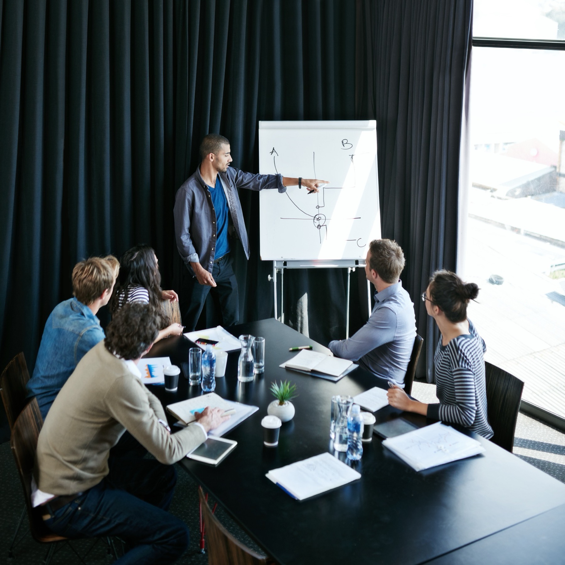 A marketing team is in a meeting. One of the consultants is presenting his findings to five of his team members, who are eagerly listening and taking notes.