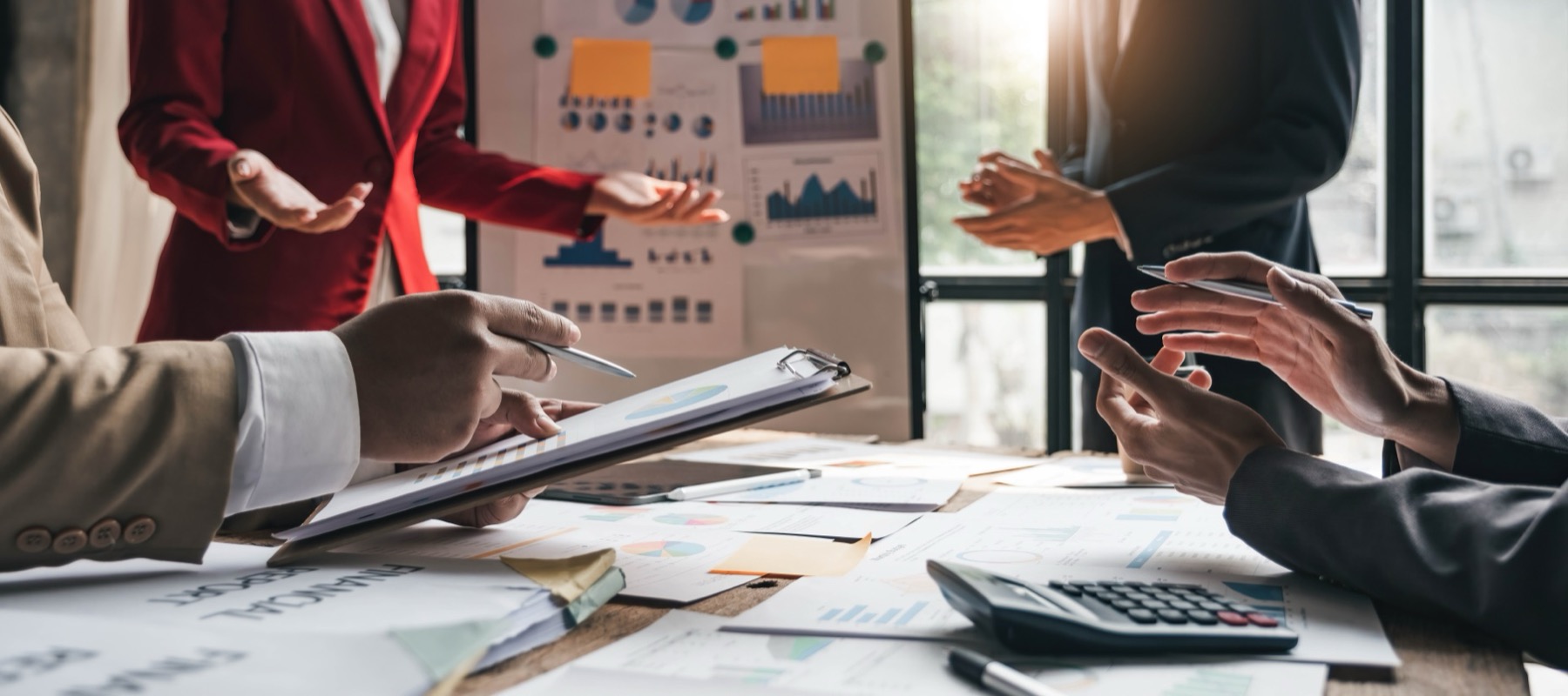A busy office filled with charts, papers, reports, and proposals pinned to the wall and laid out on a desk. Two marketers stand, discussing the latest campaign.