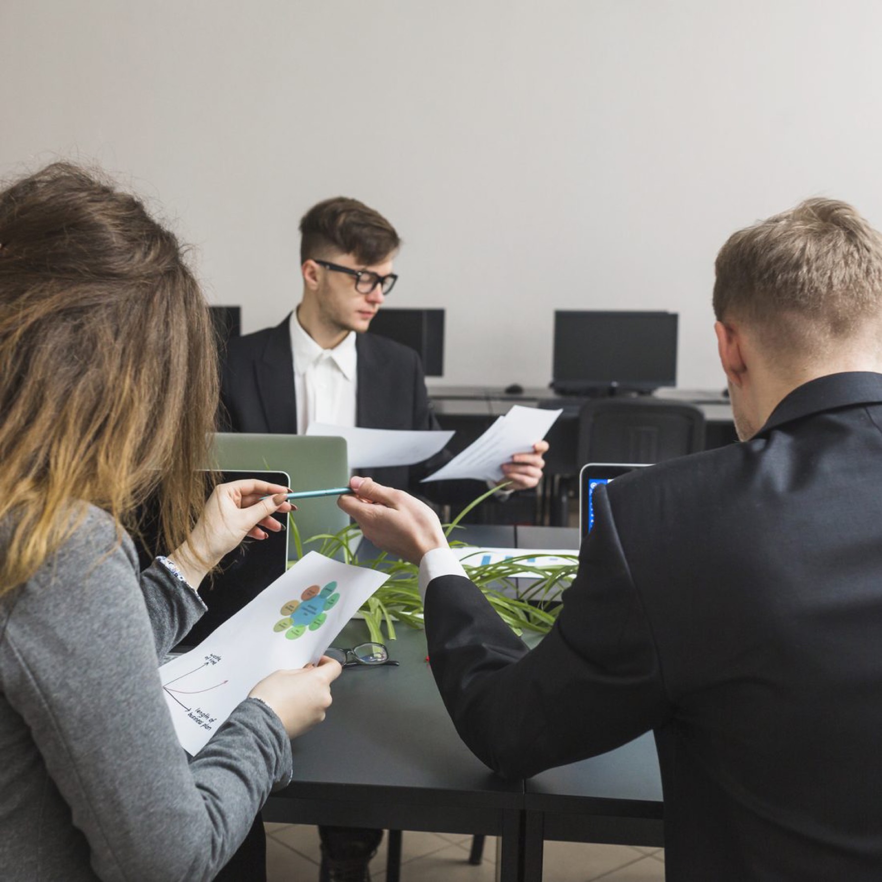 Three people sit in an open office. They are all well-dressed and are discussing the findings of their latest reports placed all over the desks.