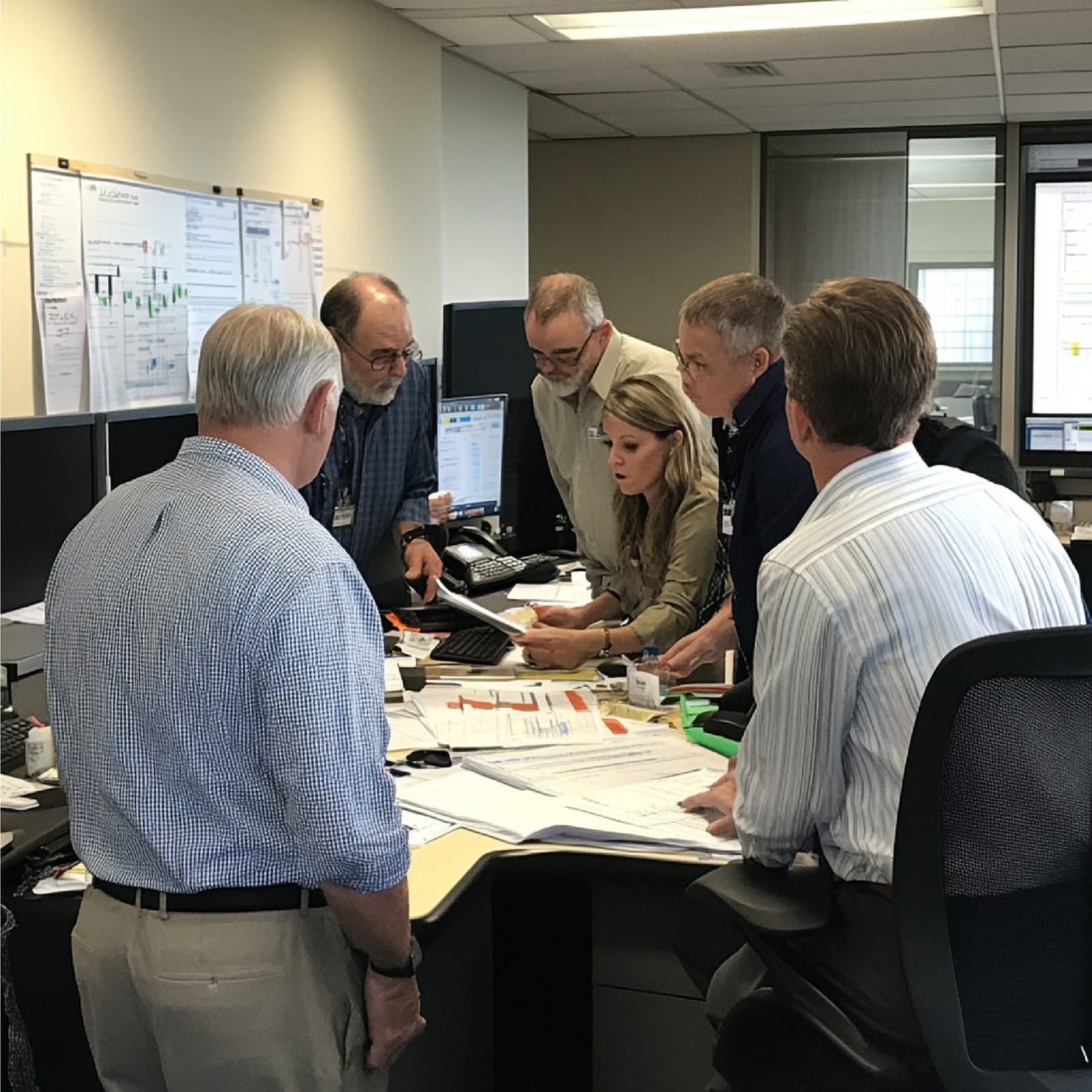 Six marketers are in a close discussion, looking over analytics and reports in their office space. Papers are all over the table surface.